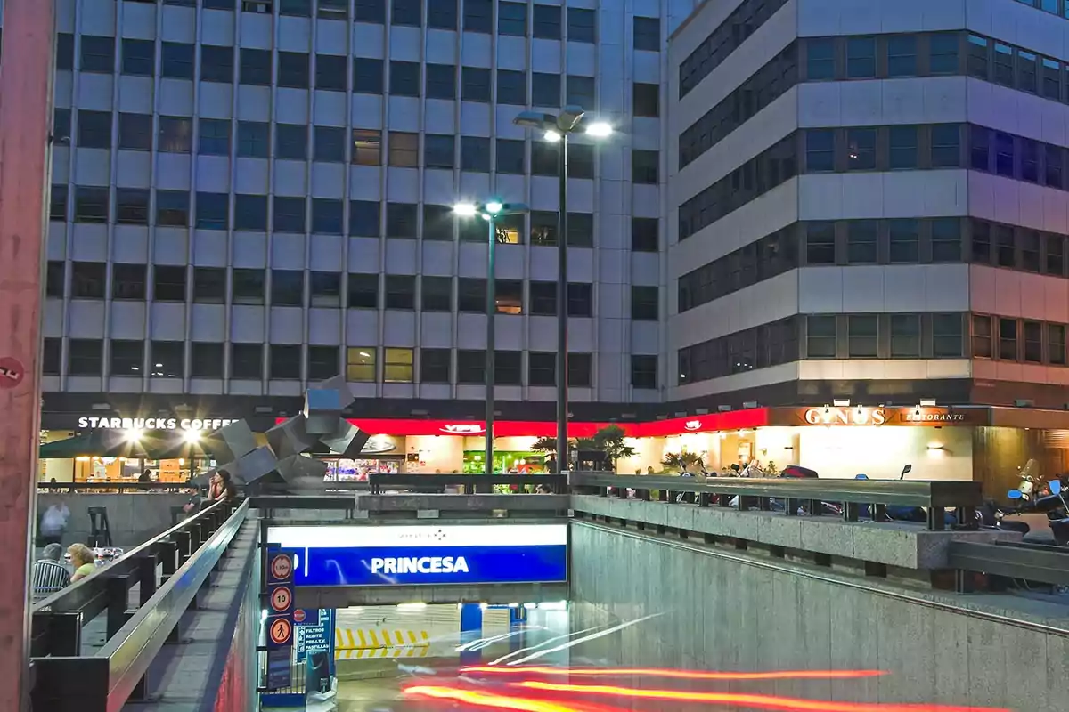 Entrada a un estacionamiento subterráneo en una zona urbana con edificios altos y varios locales comerciales iluminados.