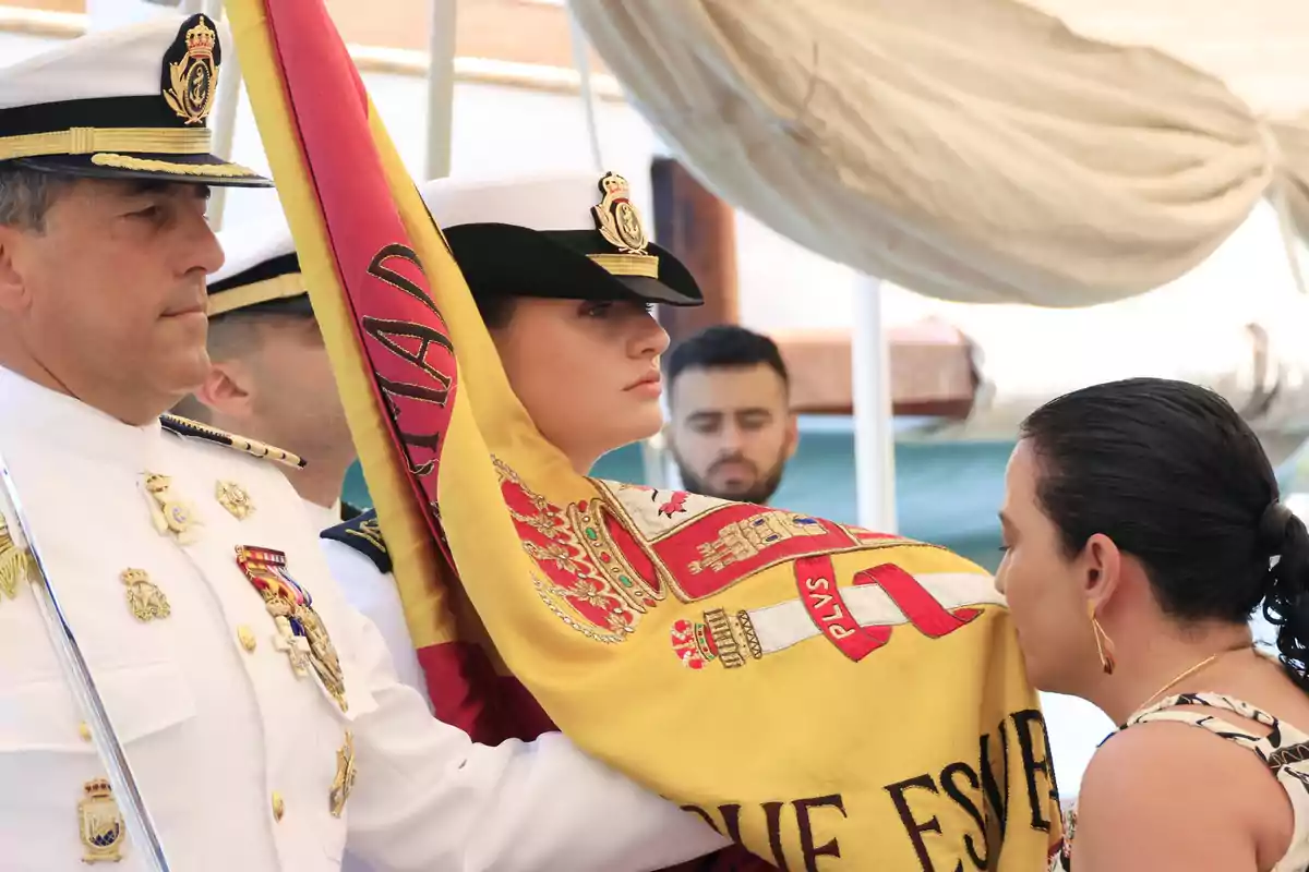 Una ceremonia militar donde una persona besa una bandera sostenida por oficiales en uniforme blanco. Una ceremonia militar donde una persona besa una bandera sostenida por oficiales en uniforme blanco.