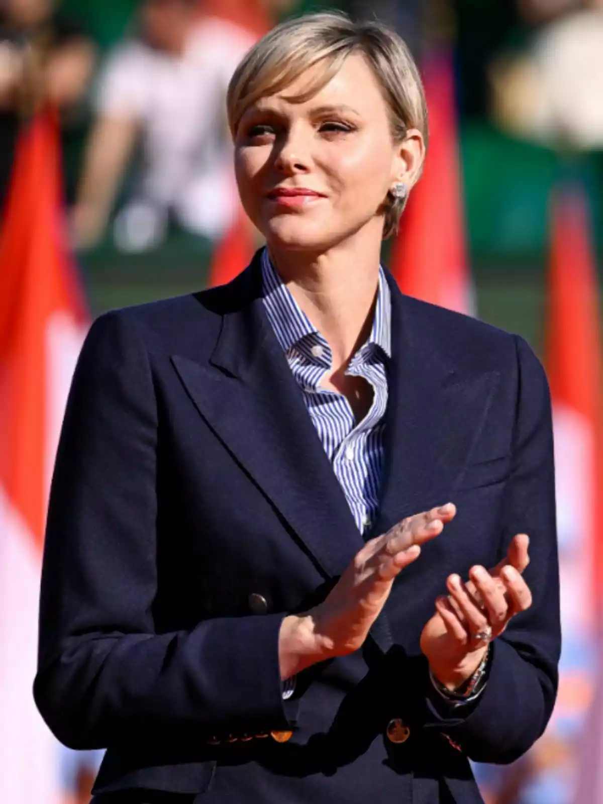 La princesa Charlene, vistiendo un traje oscuro y camisa a rayas, aplaude mientras está de pie al aire libre con banderas desenfocadas en el fondo. La princesa Charlene, vistiendo un traje oscuro y camisa a rayas, aplaude mientras está de pie al aire libre con banderas desenfocadas en el fondo.