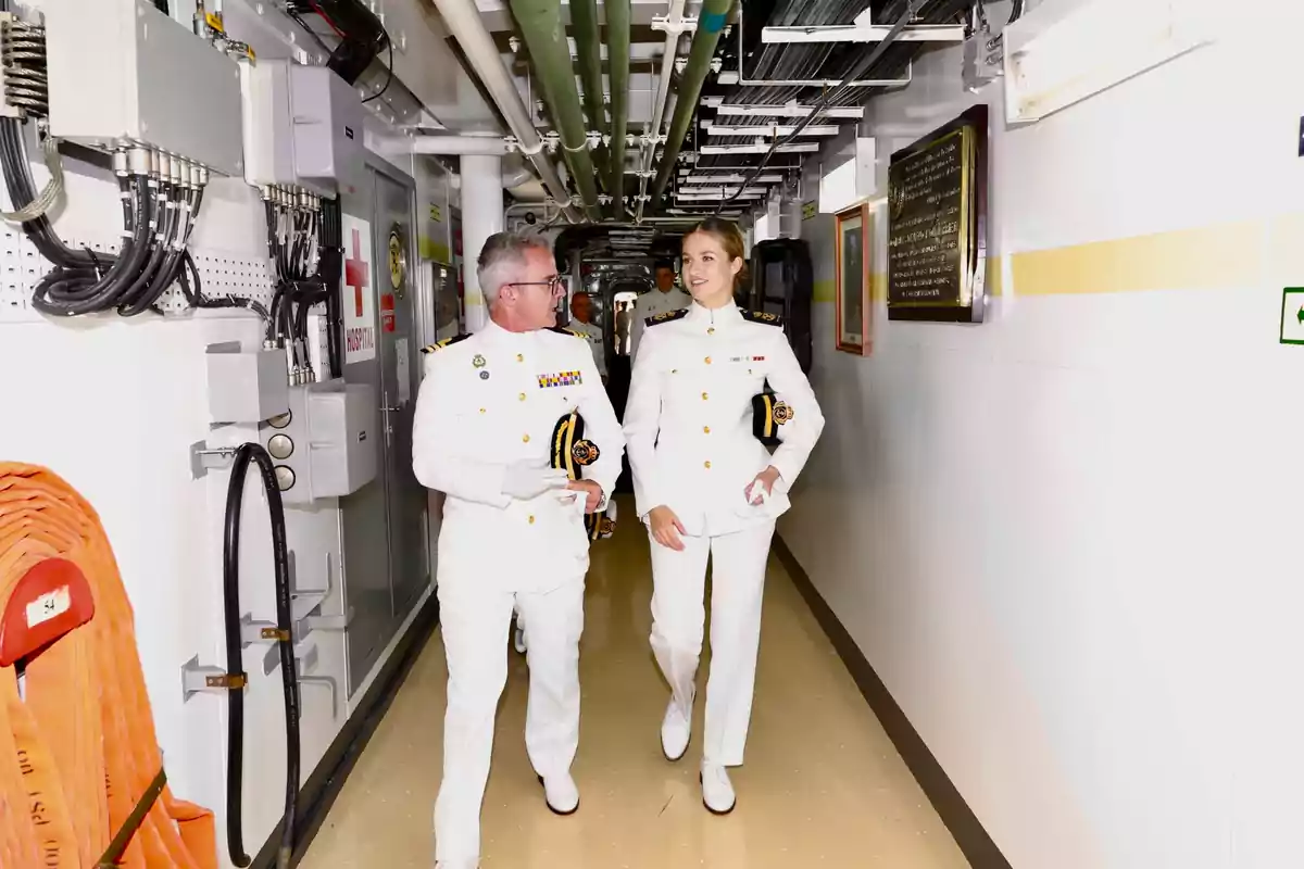 Leonor, wearing a white navy dress uniform, walks down a ship's corridor while chatting and smiling. Leonor, wearing a white navy dress uniform, walks down a ship's corridor while chatting and smiling.