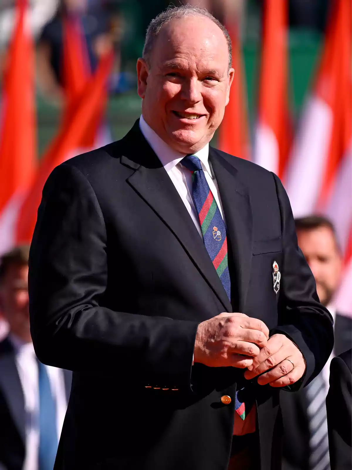 Prince Albert of Monaco in a dark suit and striped tie smiles while standing in front of several red and white flags. Prince Albert of Monaco in a dark suit and striped tie smiles while standing in front of several red and white flags.