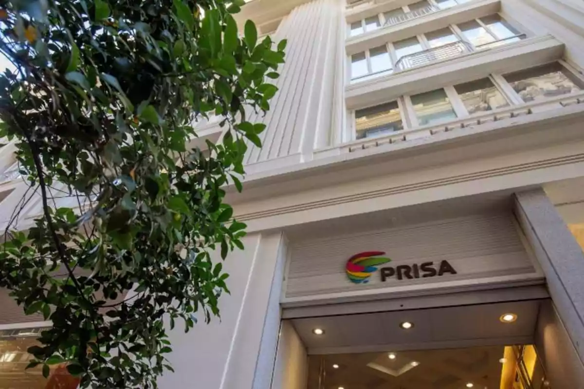 Fachada de un edificio con el logo de PRISA y un árbol en primer plano.
