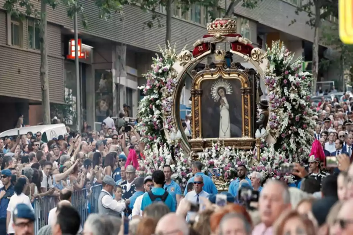 Una multitud de personas participa en una procesión religiosa en la calle, llevando una imagen sagrada adornada con flores y rodeada de devotos.