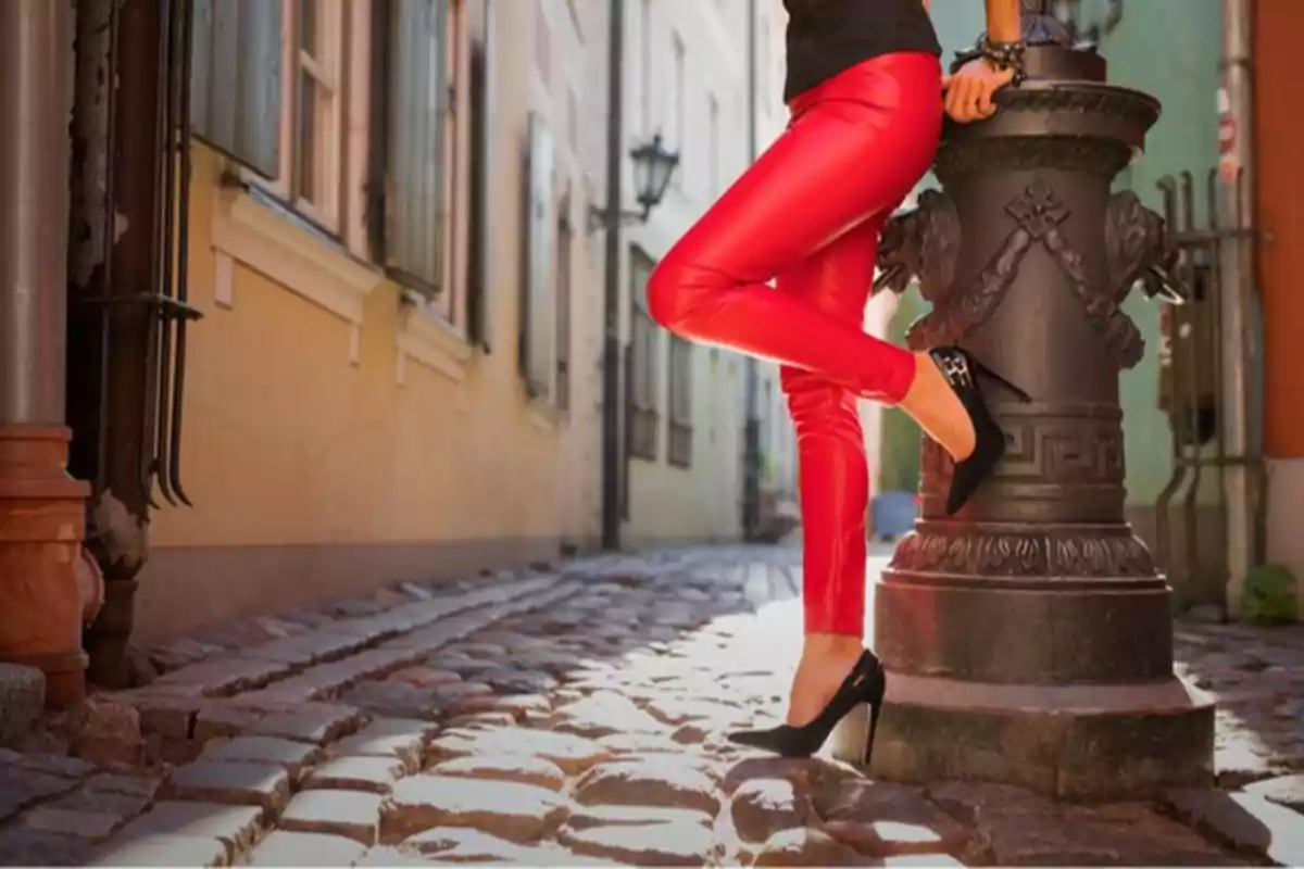 Persona con pantalones rojos y tacones negros apoyada en un poste en una calle empedrada.