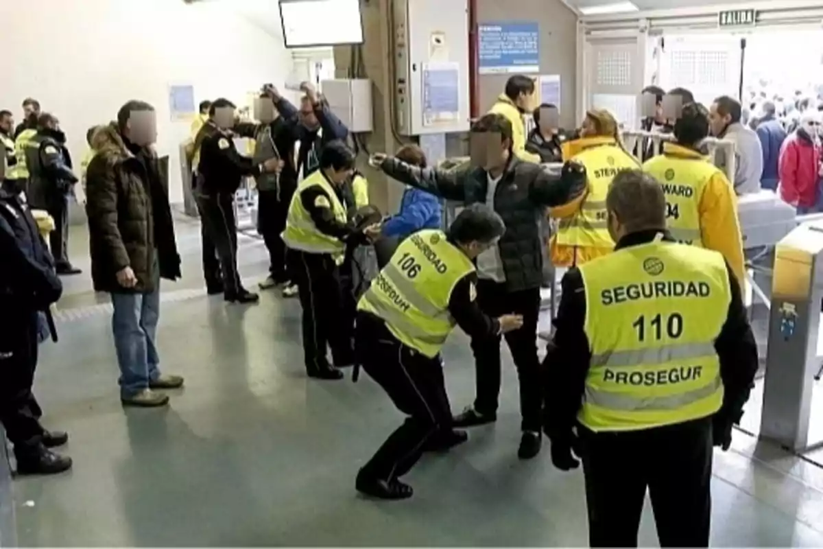 Un grupo de personas está siendo revisado por personal de seguridad en la entrada de un evento, con varios guardias vistiendo chalecos amarillos y realizando controles a los asistentes.