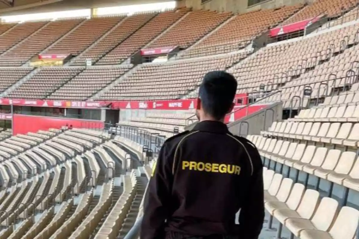 Un guardia de seguridad con uniforme de Prosegur camina por un estadio vacío con filas de asientos vacíos a su alrededor. Un guardia de seguridad con uniforme de Prosegur camina por un estadio vacío con filas de asientos vacíos a su alrededor.