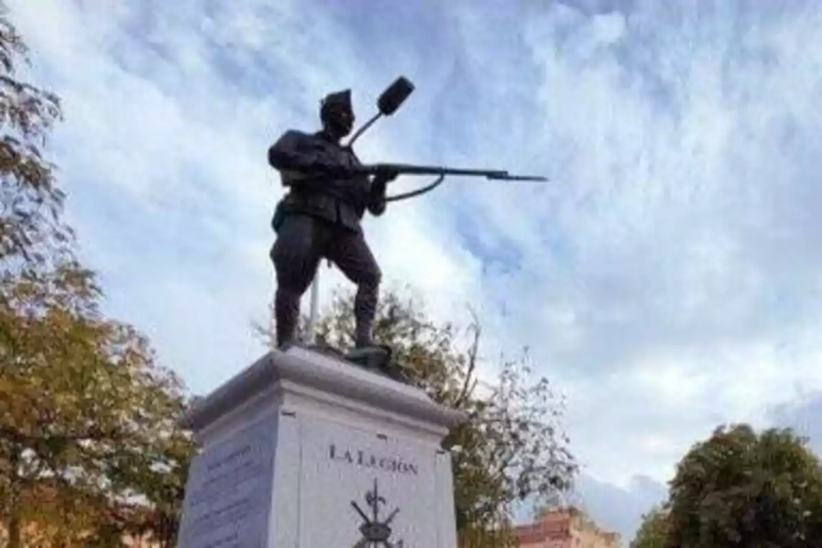 Estatua de un soldado con rifle sobre un pedestal con la inscripción "LA LEGIÓN" en un entorno al aire libre con árboles y cielo nublado.