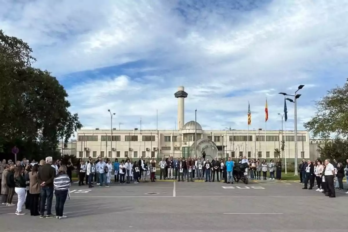 Un grupo de personas se reúne frente a un edificio con una torre y varias banderas ondeando, en un día parcialmente nublado.