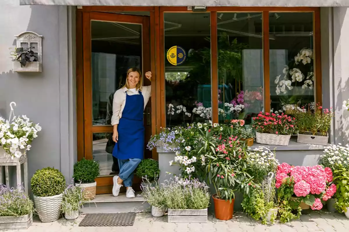Mujer con delantal azul sonriendo en la entrada de una florería rodeada de plantas y flores de colores Mujer con delantal azul sonriendo en la entrada de una florería rodeada de plantas y flores de colores
