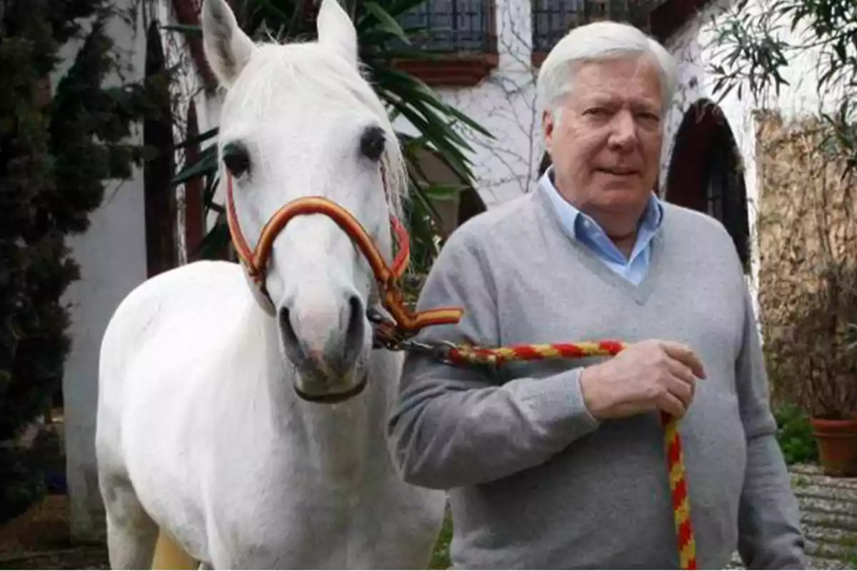 Un hombre de cabello canoso sostiene la cuerda de un caballo blanco en un entorno al aire libre.