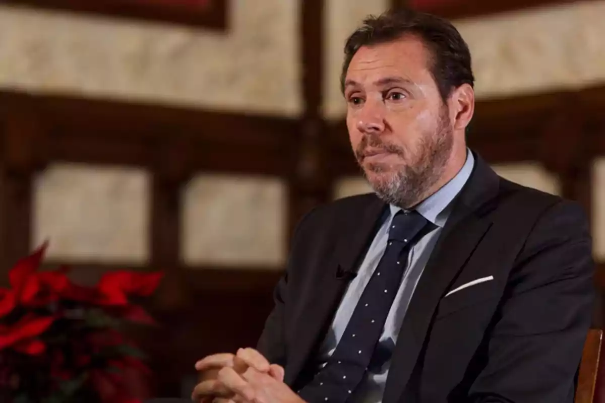 Hombre con traje oscuro y corbata azul sentado en un ambiente interior con decoración de madera y una planta roja al fondo.