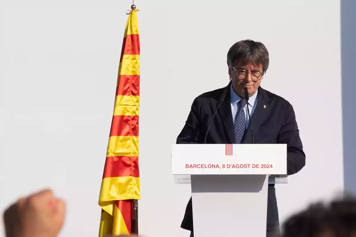 Un hombre de traje y corbata habla en un podio con un micrófono, junto a una bandera con franjas amarillas y rojas, con la inscripción 