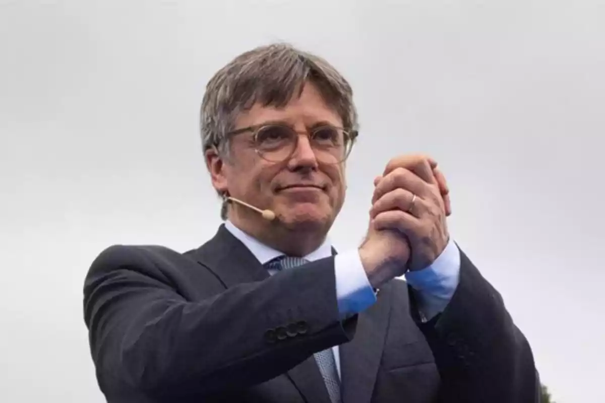 Hombre con gafas y micrófono de diadema vestido con traje oscuro y corbata, juntando las manos frente a un fondo gris