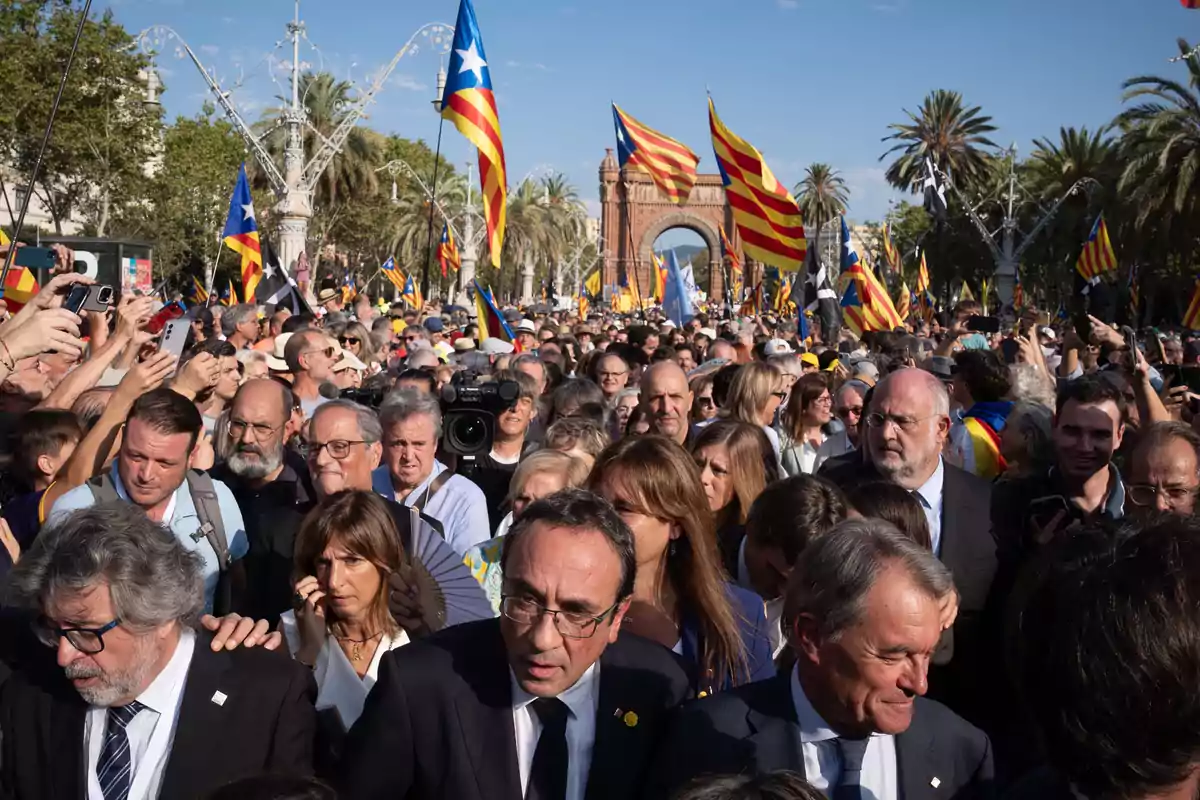 Una multitud de personas se reúne en una manifestación, ondeando banderas con los colores de Cataluña, con el Arco de Triunfo de Barcelona visible al fondo.