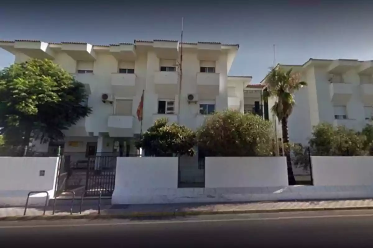 Edificio blanco de tres pisos con balcones y una bandera en la entrada, rodeado de árboles y arbustos.