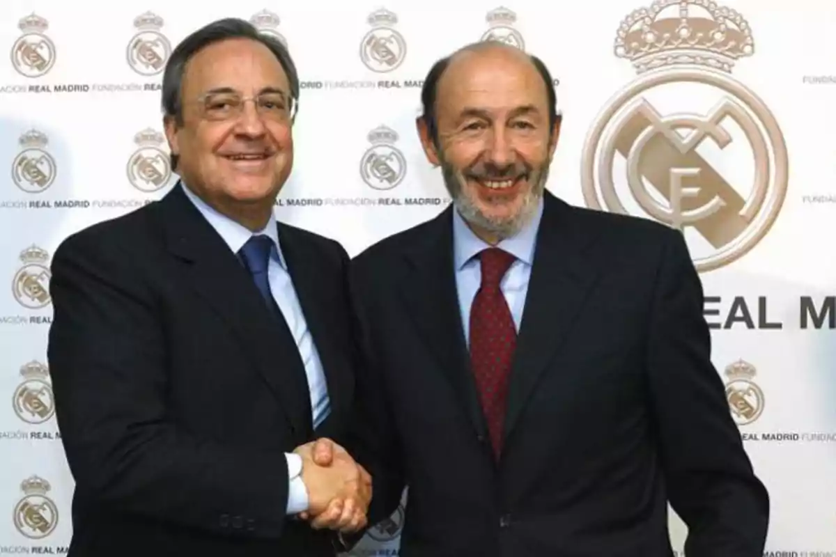 Dos hombres de traje sonríen y se dan la mano frente a un fondo con el logo del Real Madrid.