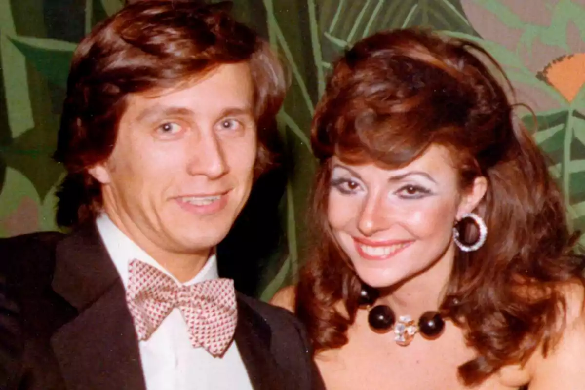 Una pareja sonriente posando para una foto, el hombre lleva un esmoquin con pajarita y la mujer lleva un collar de perlas negras y pendientes grandes.