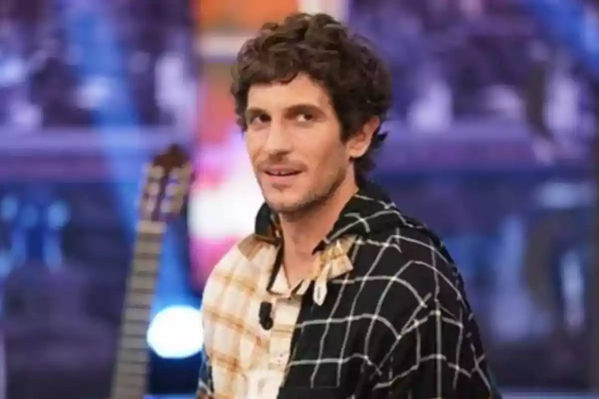 Hombre con cabello rizado y barba ligera, vistiendo una camisa de cuadros, con una guitarra en el fondo.