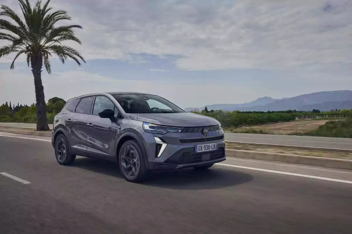 Un Renault Symbioz Hybrid gris circula por una carretera rodeada de palmeras y montañas en el fondo bajo un cielo parcialmente nublado.