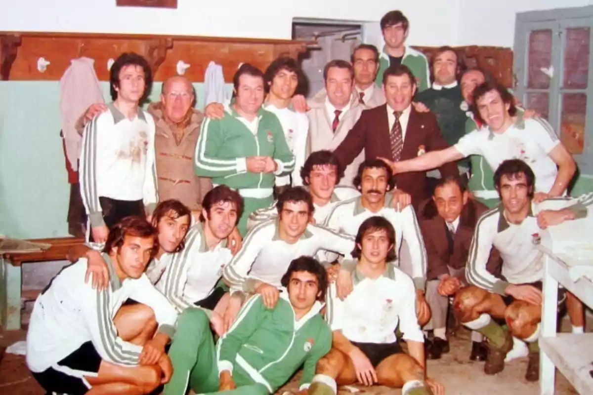 Un grupo de jugadores de fútbol y entrenadores posan juntos en un vestuario, algunos llevan uniformes deportivos y otros trajes formales, todos parecen estar celebrando o posando después de un partido.