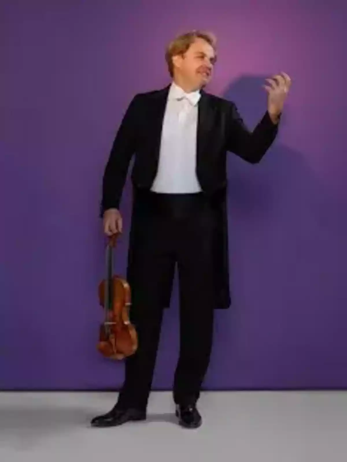 Un hombre con esmoquin sostiene un violín en una mano mientras hace un gesto con la otra frente a un fondo morado. Un hombre con esmoquin sostiene un violín en una mano mientras hace un gesto con la otra frente a un fondo morado.