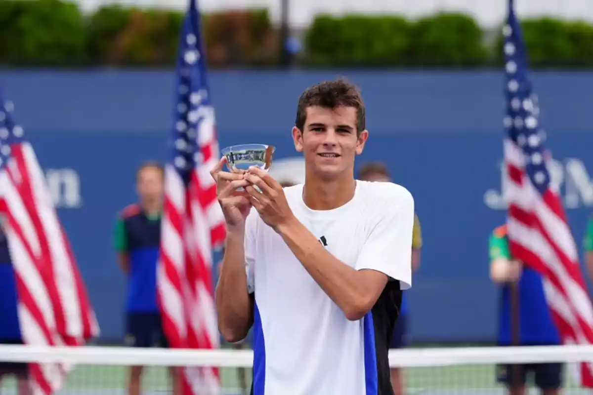 Un joven tenista sosteniendo un trofeo en una cancha de tenis con banderas estadounidenses de fondo.