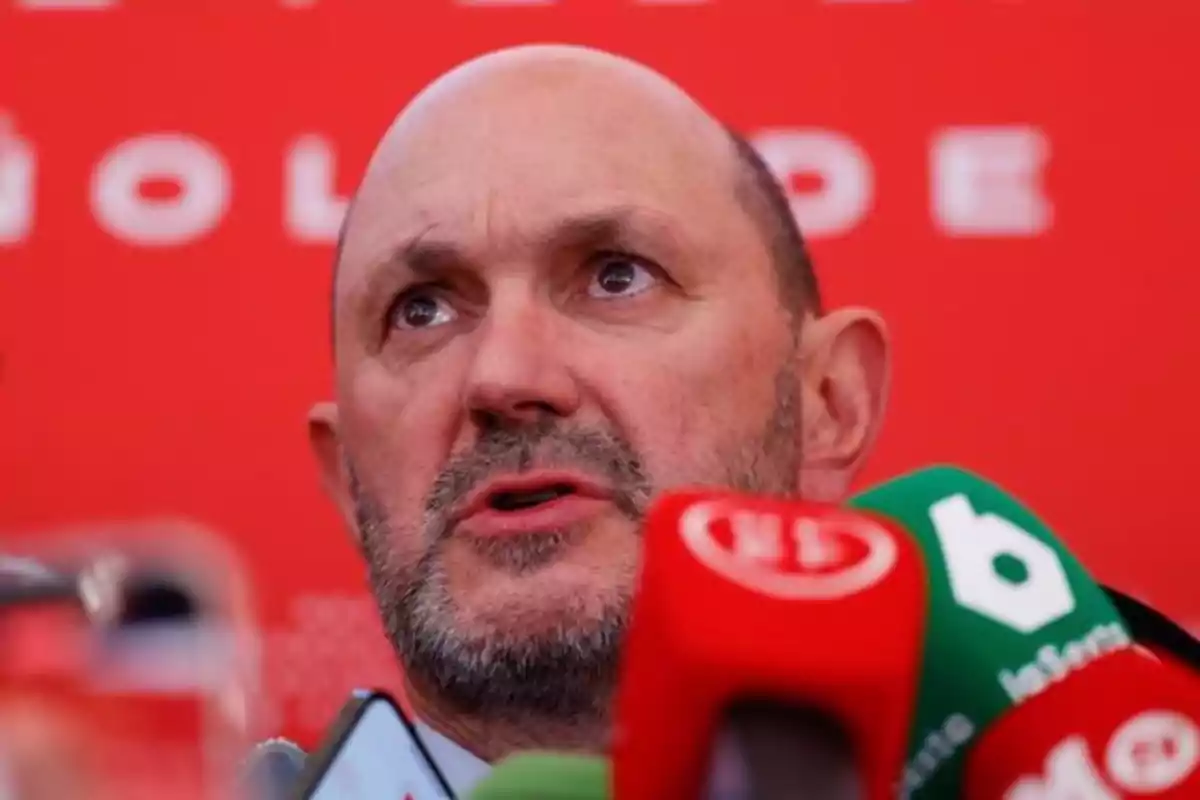 Hombre calvo hablando frente a varios micrófonos con fondo rojo.