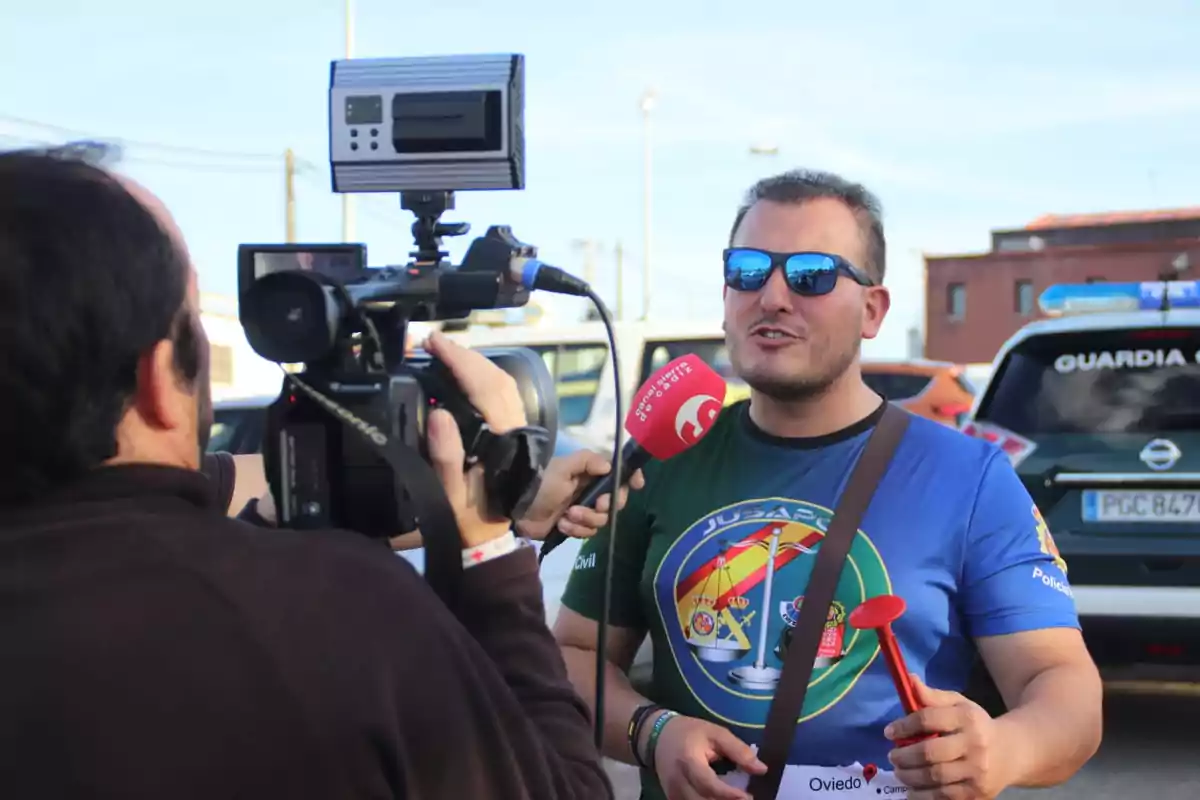 Un hombre con gafas de sol y camiseta colorida es entrevistado por un equipo de televisión en la calle mientras sostiene un objeto rojo en la mano. Un hombre con gafas de sol y camiseta colorida es entrevistado por un equipo de televisión en la calle mientras sostiene un objeto rojo en la mano.