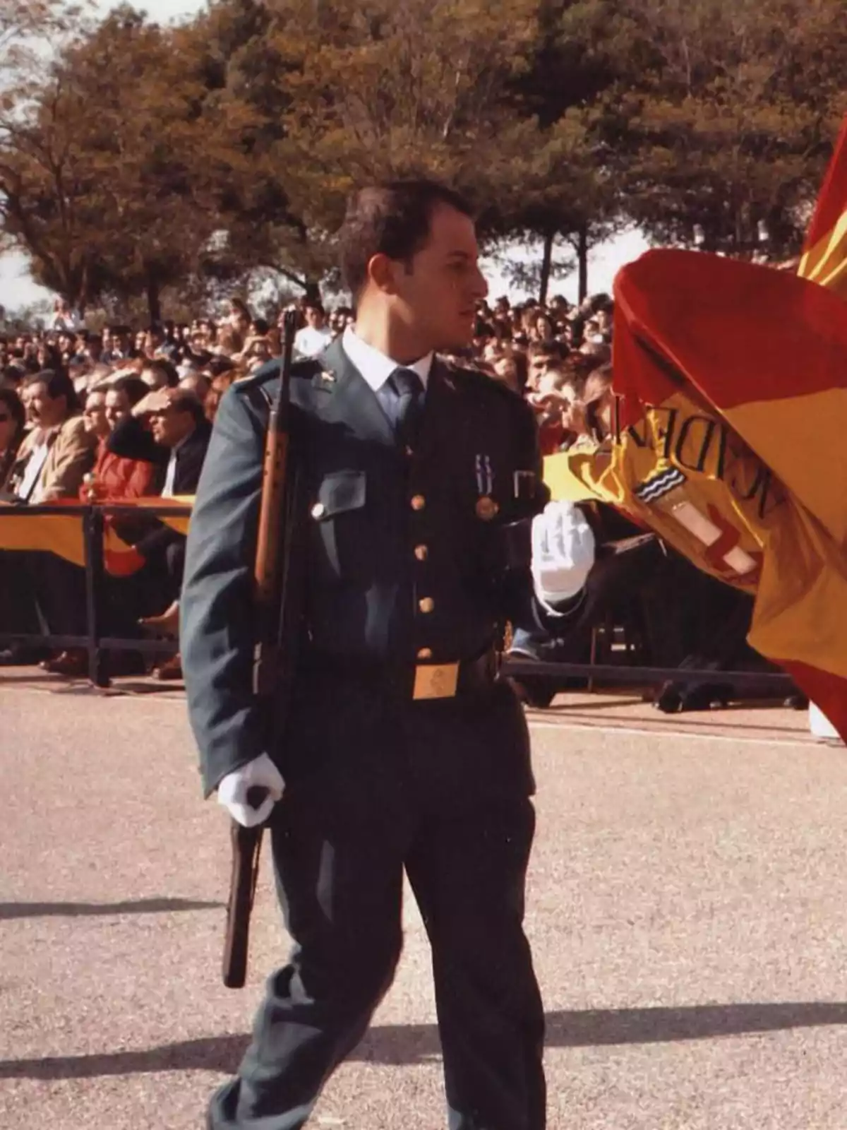 Un miembro de la Guardia Civil en uniforme formal sostiene una bandera durante un evento al aire libre con una multitud de personas y árboles al fondo. Un miembro de la Guardia Civil en uniforme formal sostiene una bandera durante un evento al aire libre con una multitud de personas y árboles al fondo.