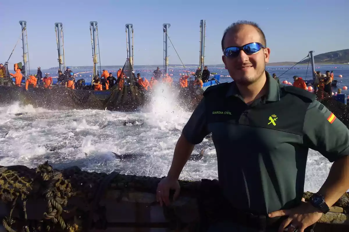 Un hombre con uniforme de la Guardia Civil posa frente a un grupo de personas vestidas de naranja que trabajan en una estructura sobre el agua, con olas rompiendo alrededor. Un hombre con uniforme de la Guardia Civil posa frente a un grupo de personas vestidas de naranja que trabajan en una estructura sobre el agua, con olas rompiendo alrededor.
