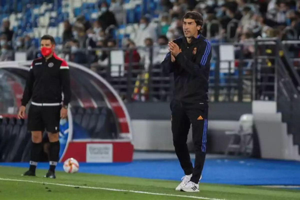 Un entrenador de fútbol aplaude en la línea lateral del campo mientras otro hombre con mascarilla roja observa, con un estadio lleno de espectadores al fondo. Un entrenador de fútbol aplaude en la línea lateral del campo mientras otro hombre con mascarilla roja observa, con un estadio lleno de espectadores al fondo.