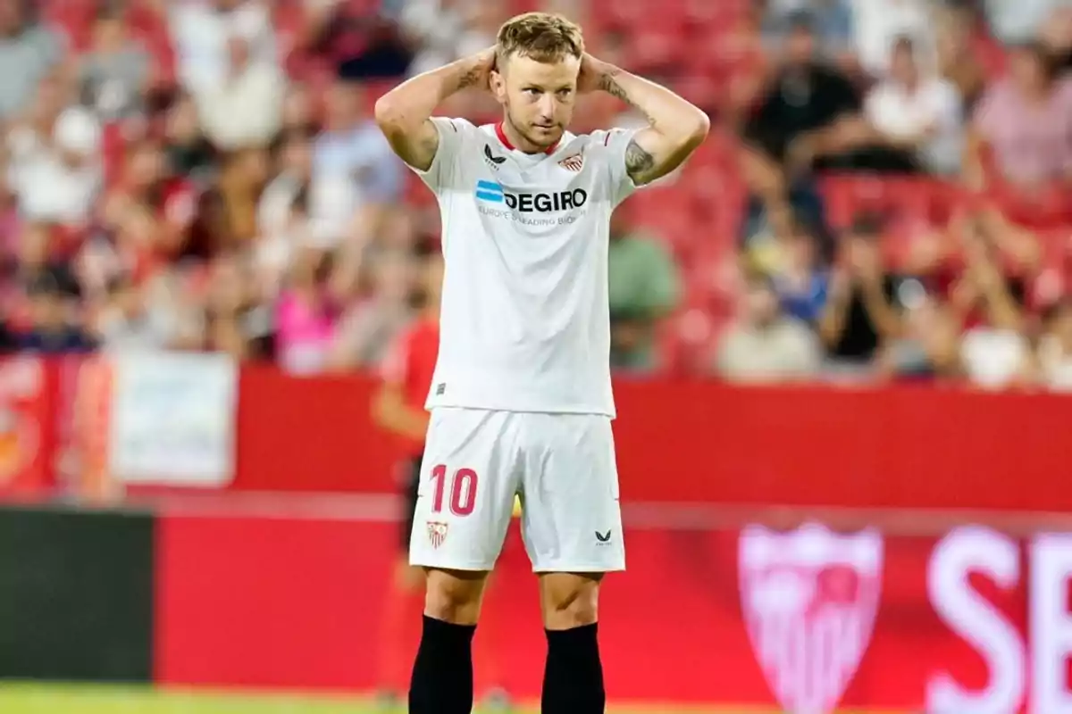 Futbolista del Sevilla con el número 10 en el uniforme blanco, de pie en el campo con las manos en la cabeza y fondo de gradas llenas. Futbolista del Sevilla con el número 10 en el uniforme blanco, de pie en el campo con las manos en la cabeza y fondo de gradas llenas.