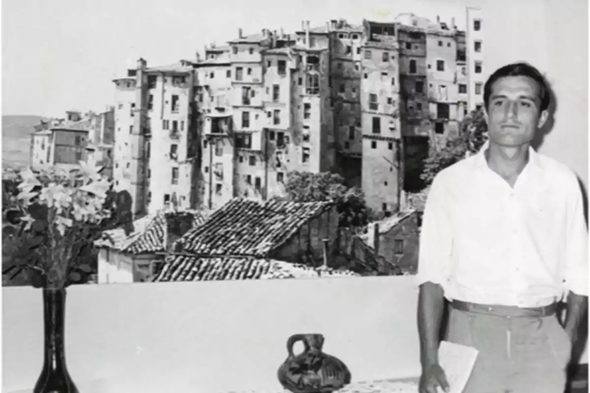 Un hombre de pie junto a una mesa con un jarrón de flores y una jarra, con un fondo de edificios antiguos.