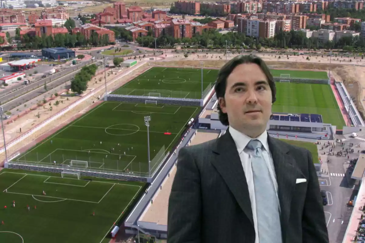 Un hombre con traje frente a un complejo deportivo con campos de fútbol y edificios residenciales al fondo.