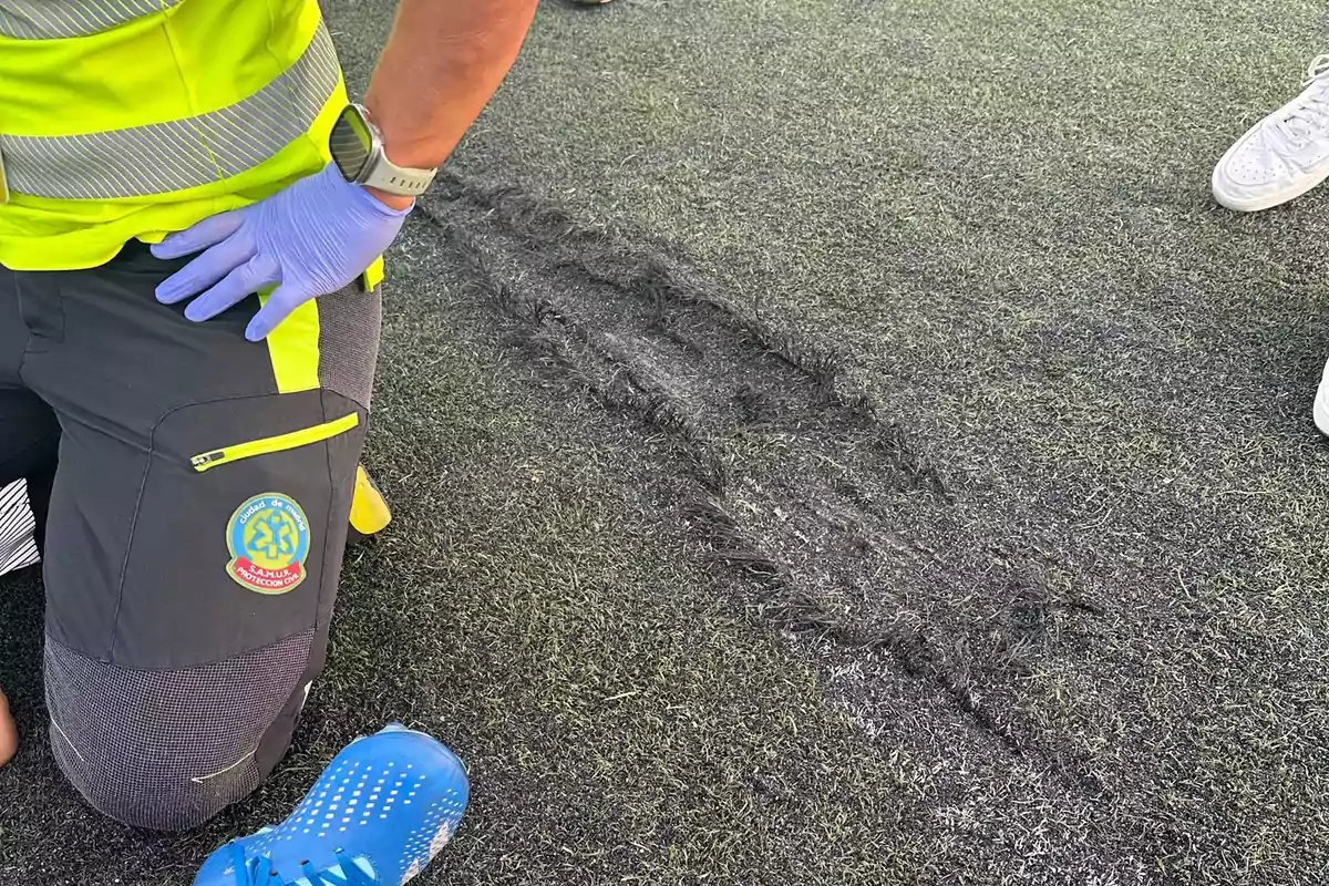 Persona con uniforme de protección civil arrodillada en un campo de césped artificial con marcas de quemaduras.