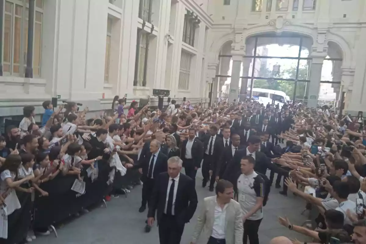 Un grupo de personas vestidas de traje camina por un pasillo mientras una multitud de aficionados los saluda y toma fotos.