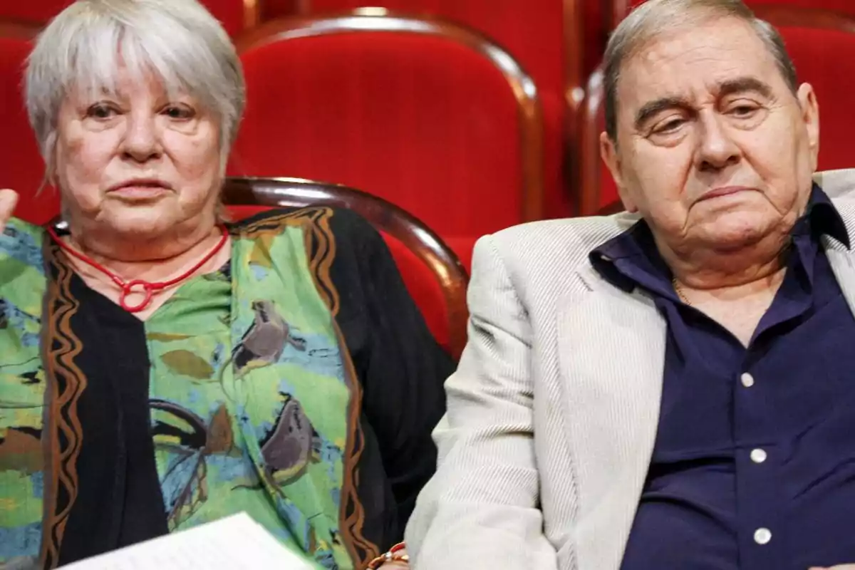 Dos personas mayores sentadas en sillas rojas, una mujer con cabello canoso y un hombre con chaqueta clara. Dos personas mayores sentadas en sillas rojas, una mujer con cabello canoso y un hombre con chaqueta clara.