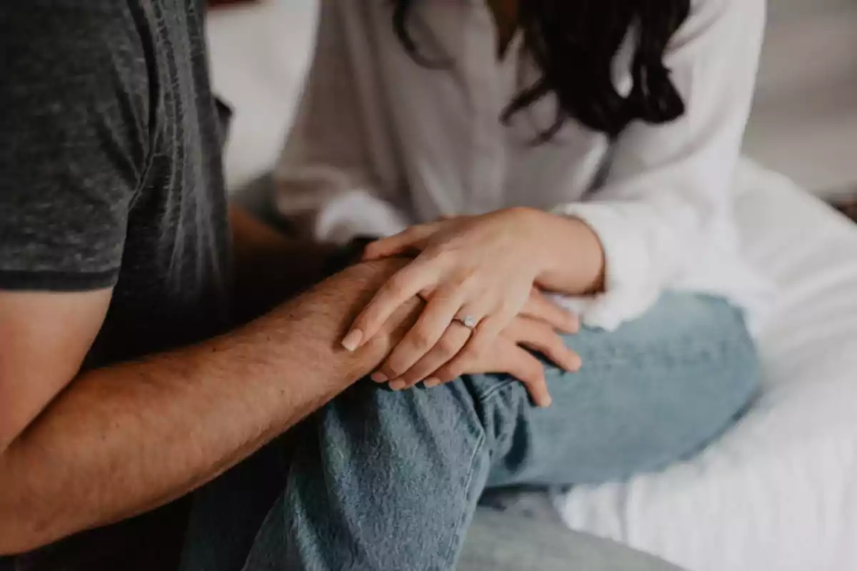 Una pareja sentada en una cama, con las manos entrelazadas y una mujer mostrando un anillo de compromiso.