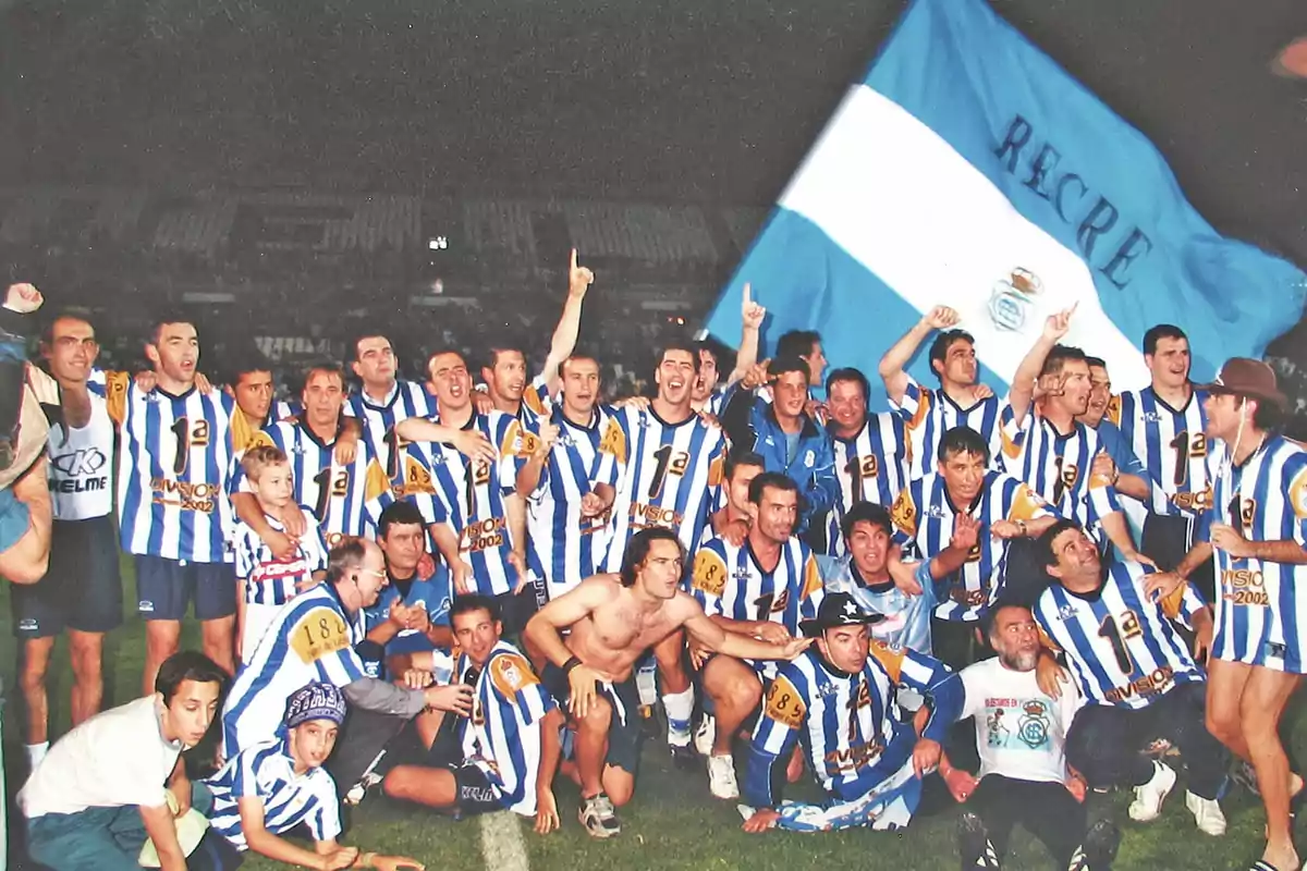 Un equipo de fútbol celebra en el campo con camisetas a rayas azules y blancas mientras sostienen una gran bandera azul y blanca.