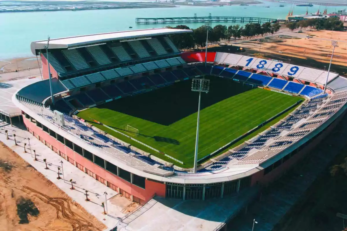 Vista aérea de un estadio de fútbol con gradas azules y blancas y el número 1889 en las gradas, ubicado cerca de un cuerpo de agua.