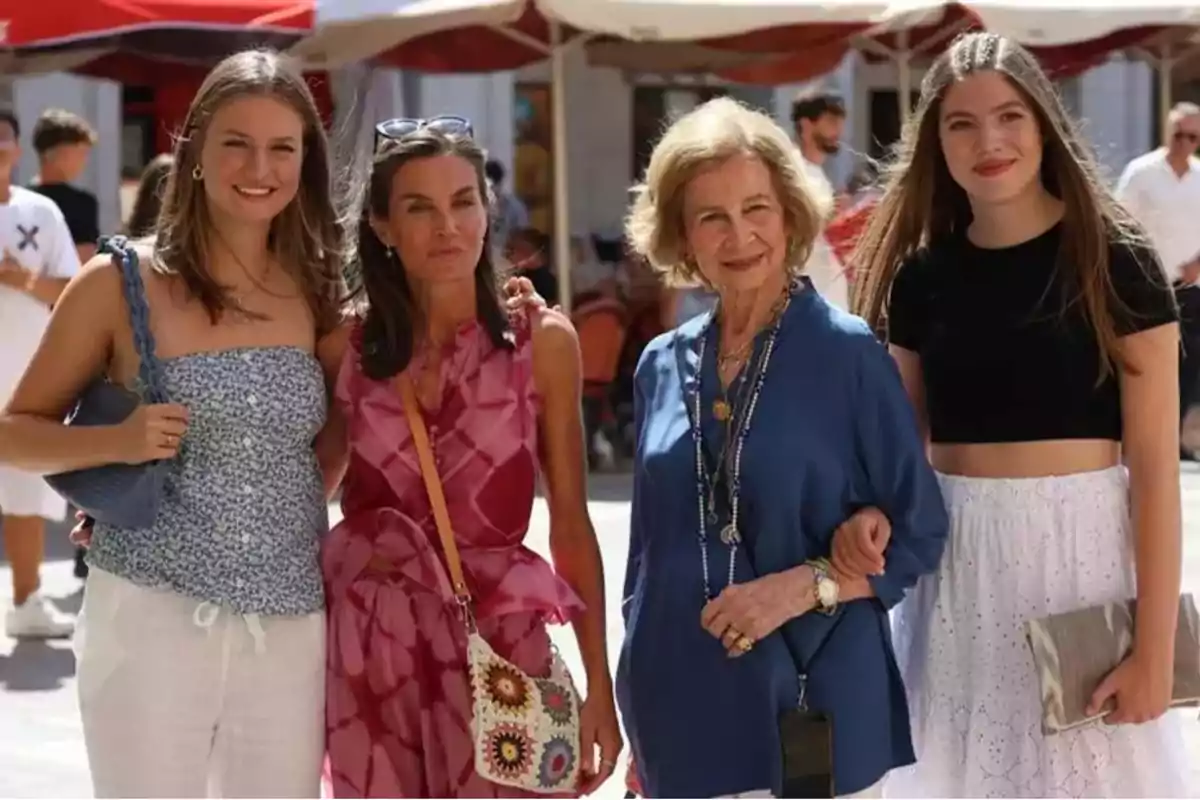 Un grupo de cuatro mujeres posando juntas al aire libre en un día soleado, con sombrillas y personas de fondo.