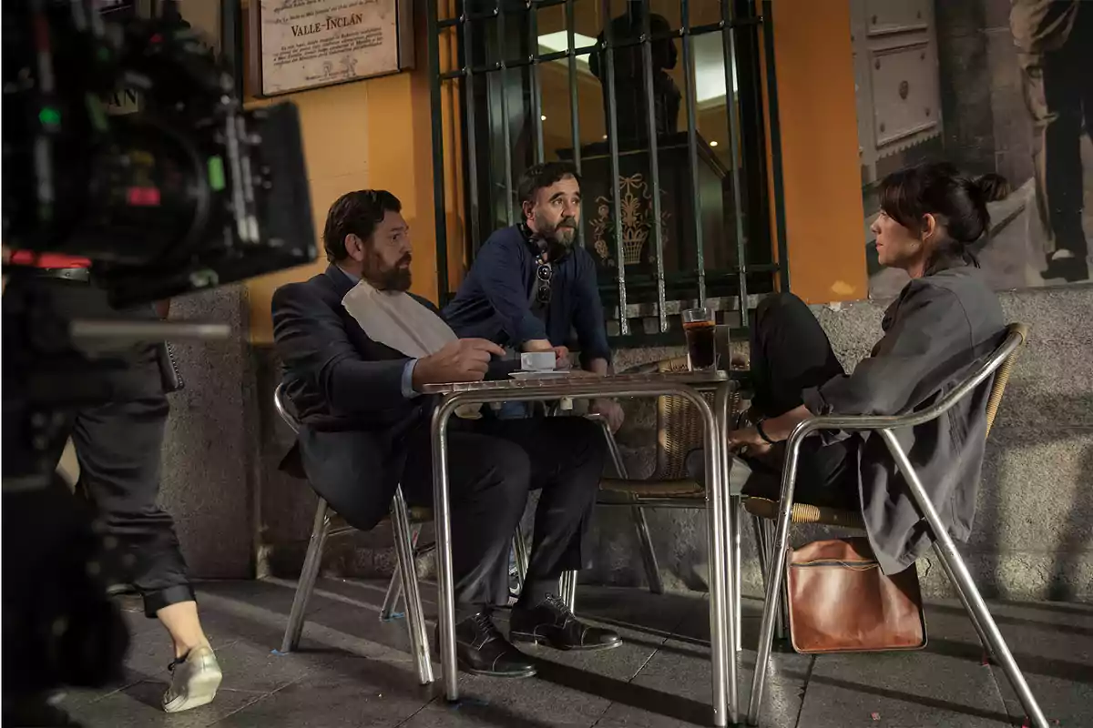 Tres personas conversan en una mesa al aire libre en un café, con una cámara de filmación visible en primer plano.