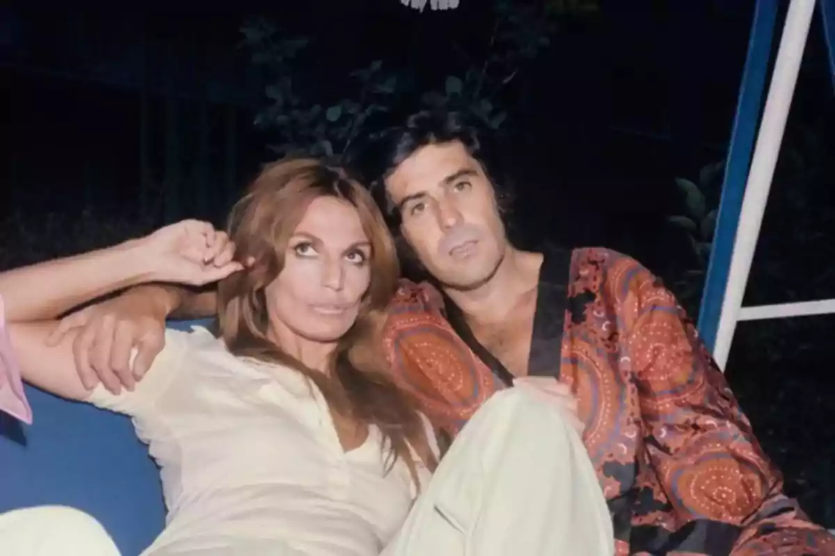 Pareja sentada en un sofá al aire libre durante la noche ambos con expresión relajada y ropa de estilo retro Pareja sentada en un sofá al aire libre durante la noche ambos con expresión relajada y ropa de estilo retro