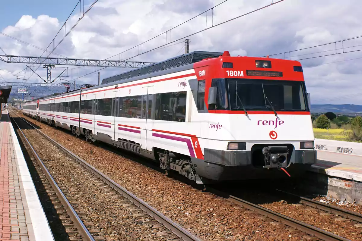 Un tren de la compañía Renfe en una estación, con un cielo parcialmente nublado al fondo.