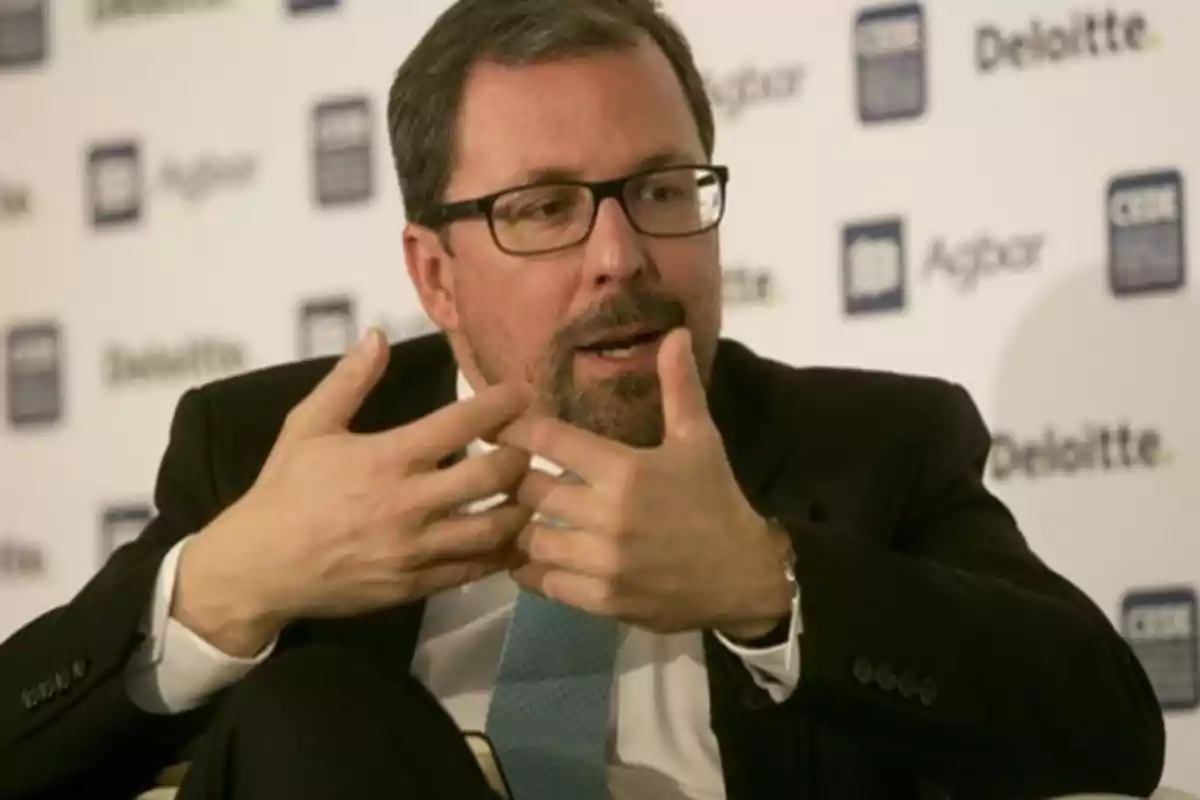 Hombre con gafas y barba hablando en un evento con logos de Deloitte y otras empresas en el fondo.