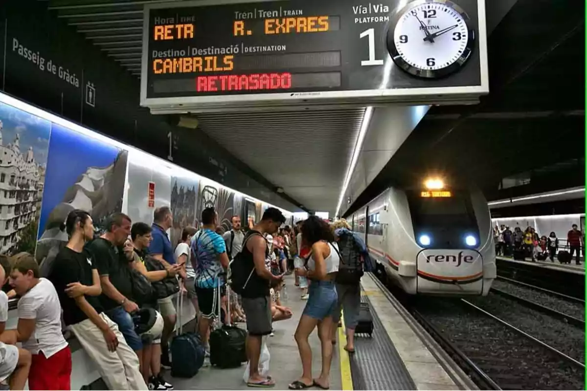 Estación de tren con muchas personas esperando en el andén, un tren de Renfe detenido y un cartel que indica que el tren con destino a Cambrils está retrasado.