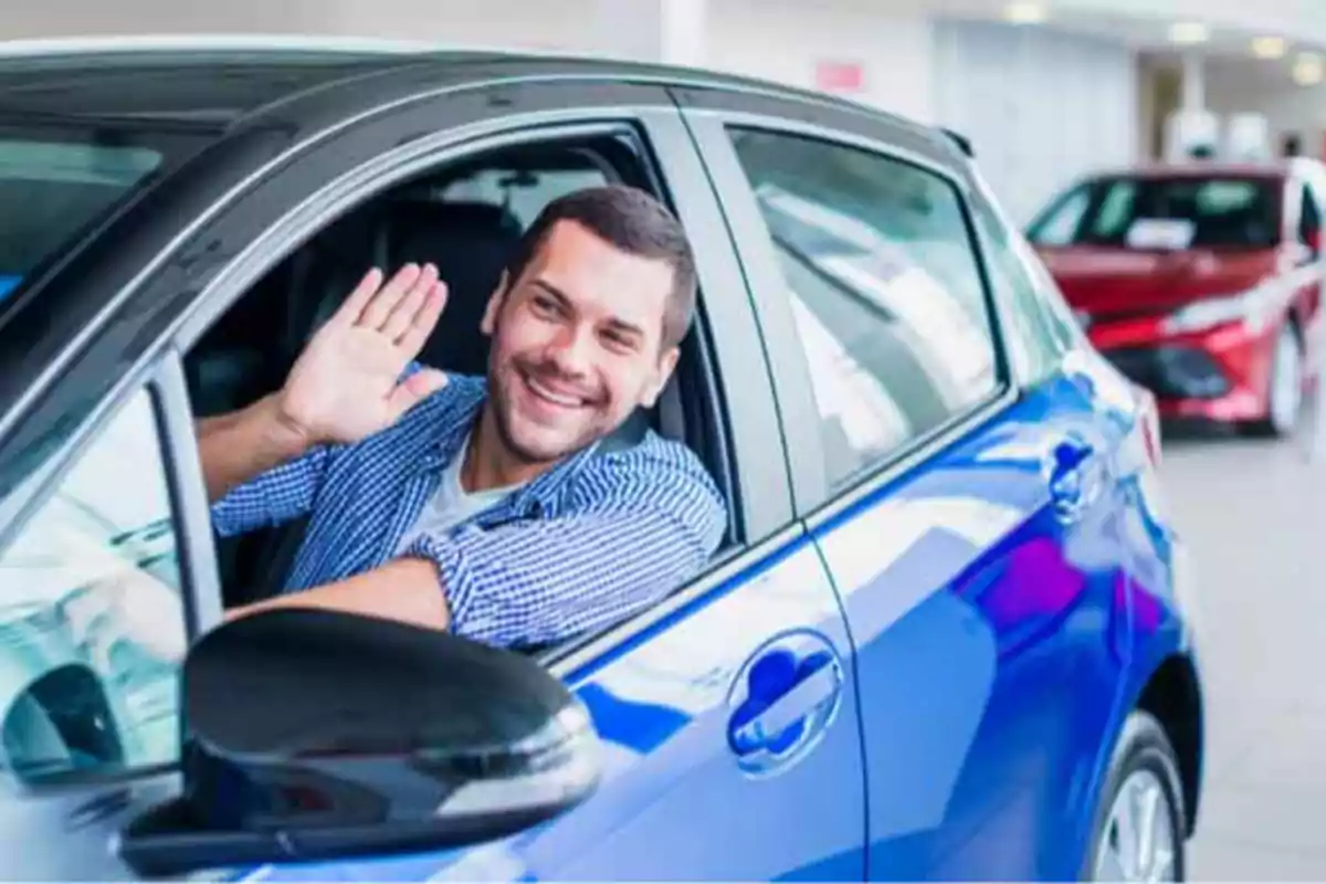Hombre sonriendo y saludando desde el interior de un coche azul en un concesionario de automóviles.