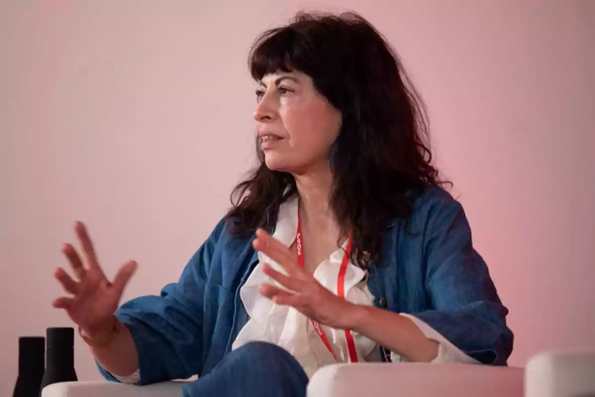 Mujer de cabello oscuro y ondulado sentada en un sillón blanco gesticulando con las manos mientras habla, lleva una chaqueta azul y una blusa blanca