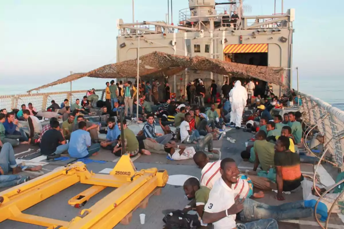 Personas sentadas y acostadas en la cubierta de un barco, algunas bajo una lona, con equipo de carga amarillo en primer plano y el mar de fondo.