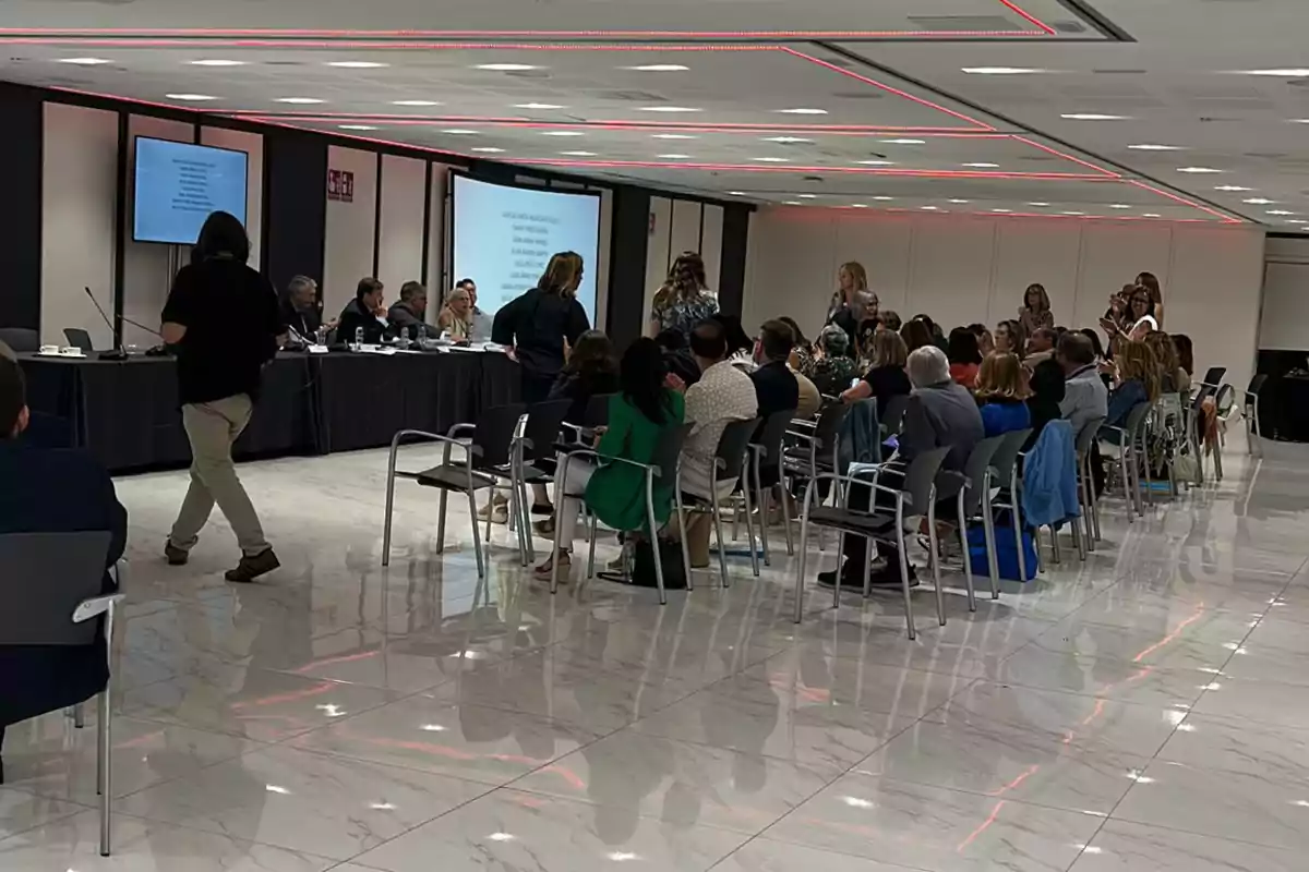 Personas asistiendo a una conferencia en una sala moderna con pantallas y luces en el techo.